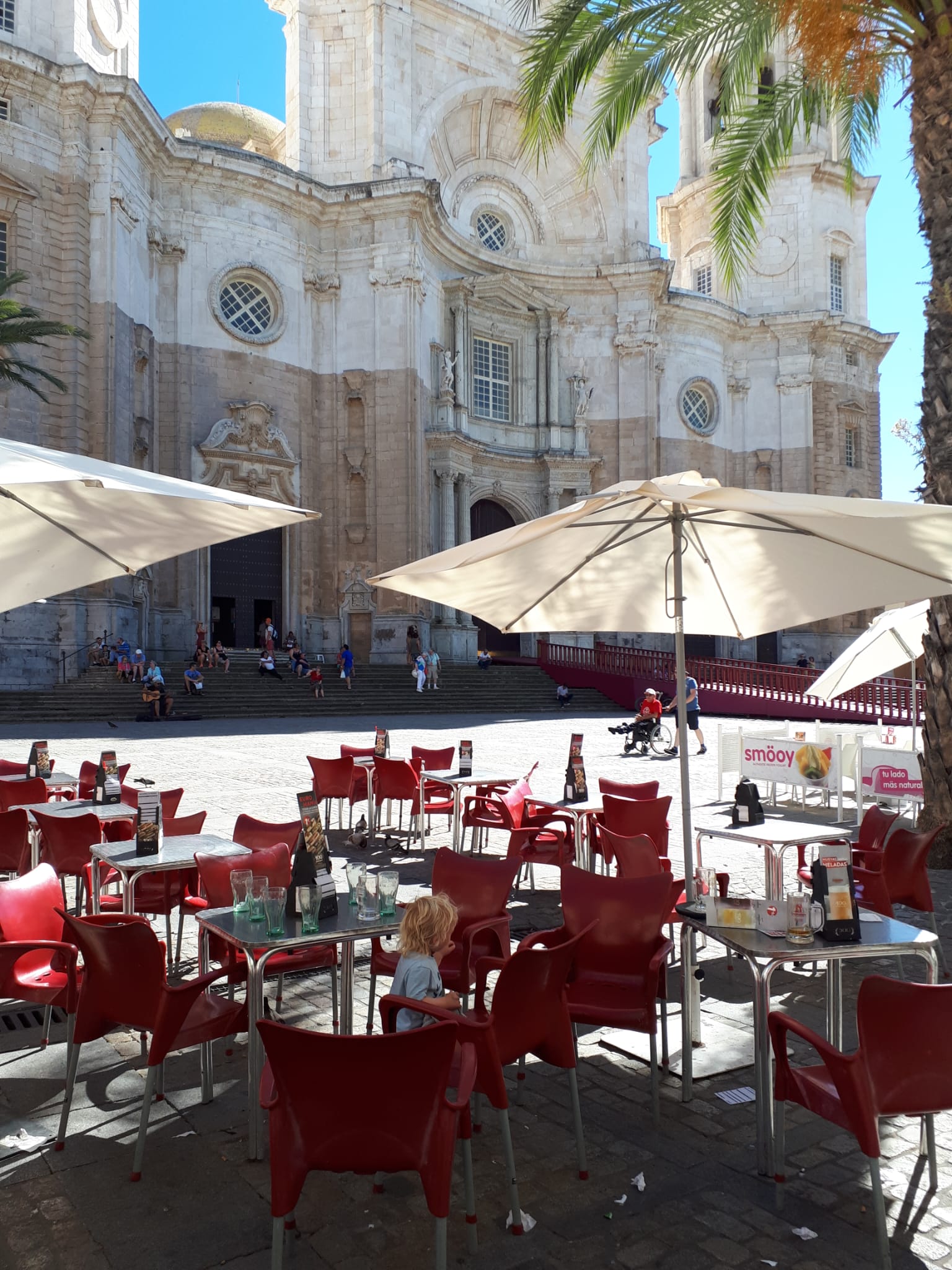 A child sitting at a café terrace with red chairs in front of a sun-drenched cathedral